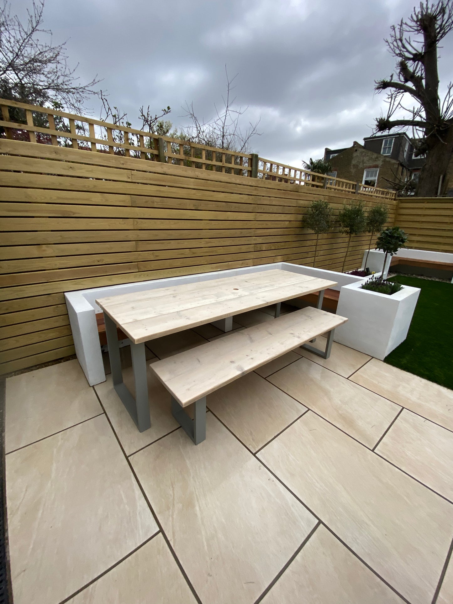 Patio area with wooden table and benches on a tiled patio, surrounded by a wooden fence and plants.