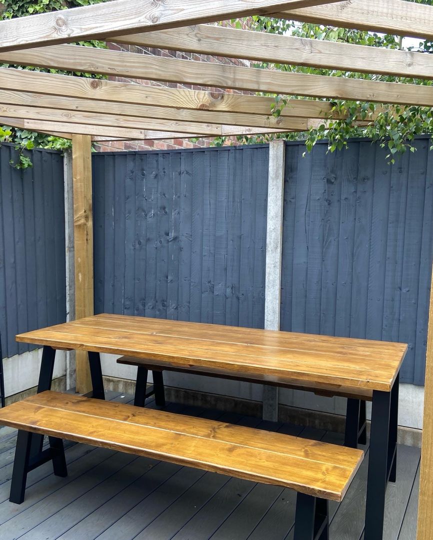 Wooden outdoor table and bench set under a wooden pergola against a gray fence.