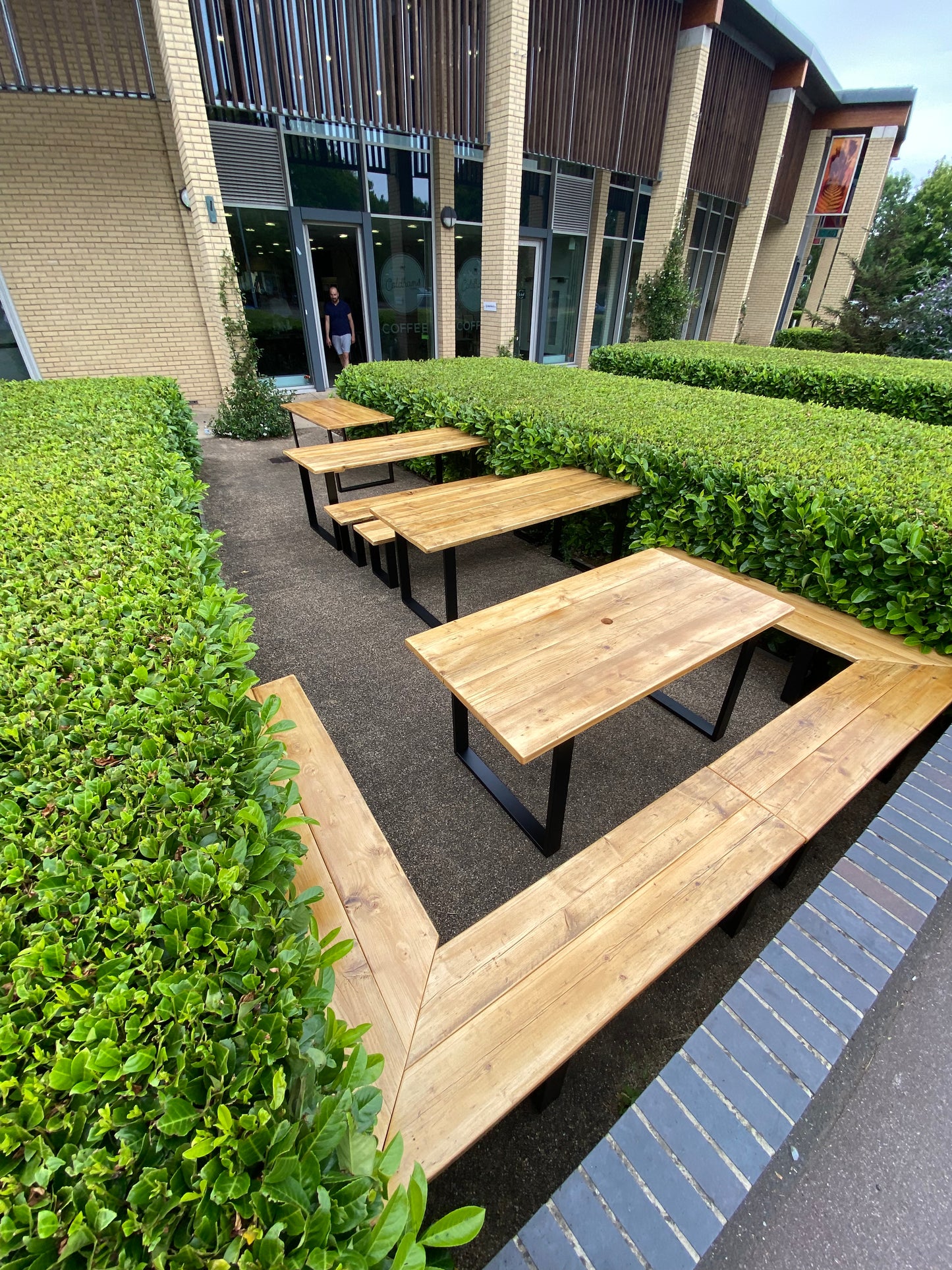 Wooden picnic tables arranged in a row with green hedges and a building in the background.